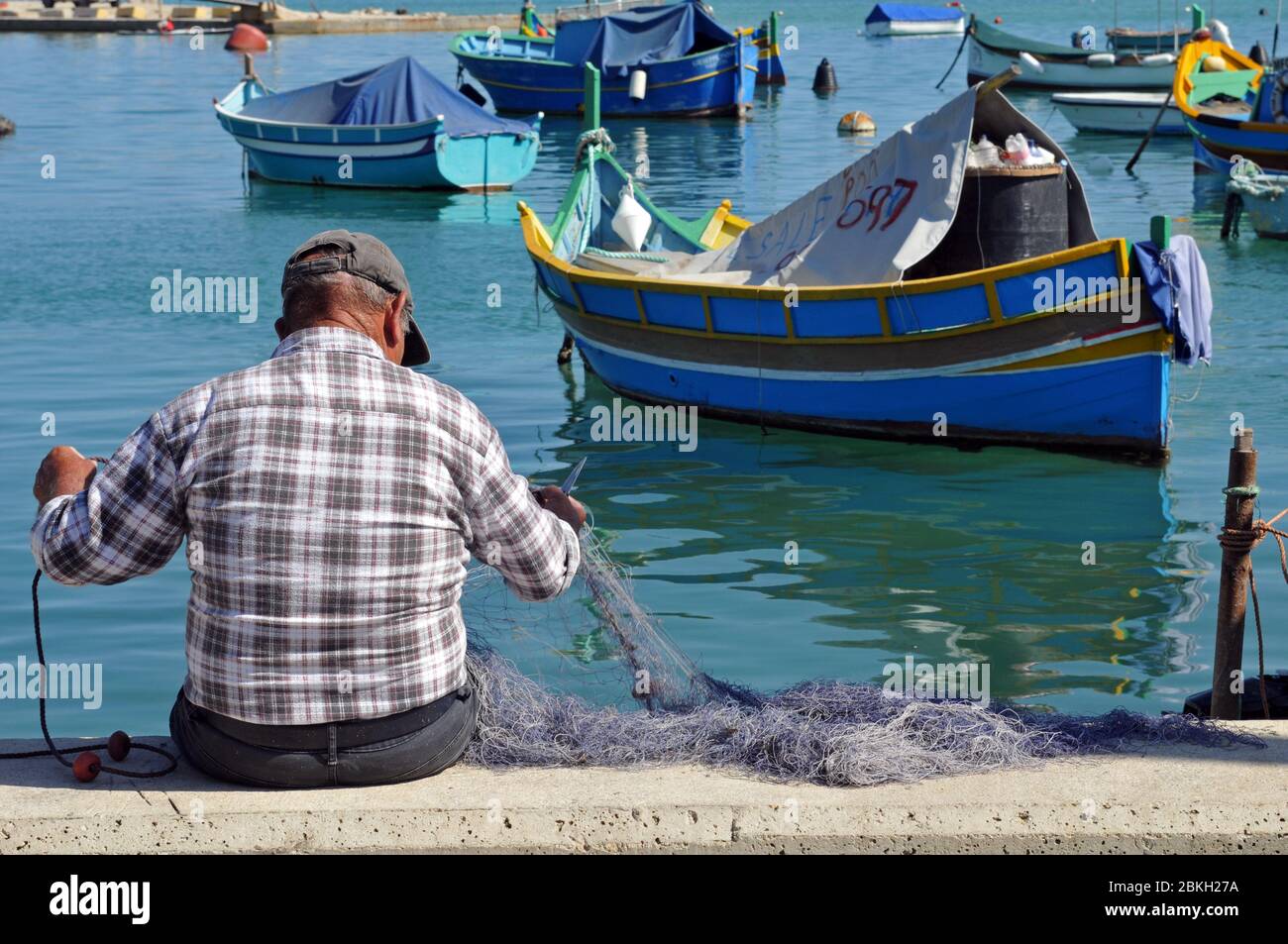 Ein Mann sitzt am Wasser und repariert ein Fischernetz im Dorf Marsaxlokk, Malta. Traditionelle maltesische Boote sind vor der Küste verankert. Stockfoto