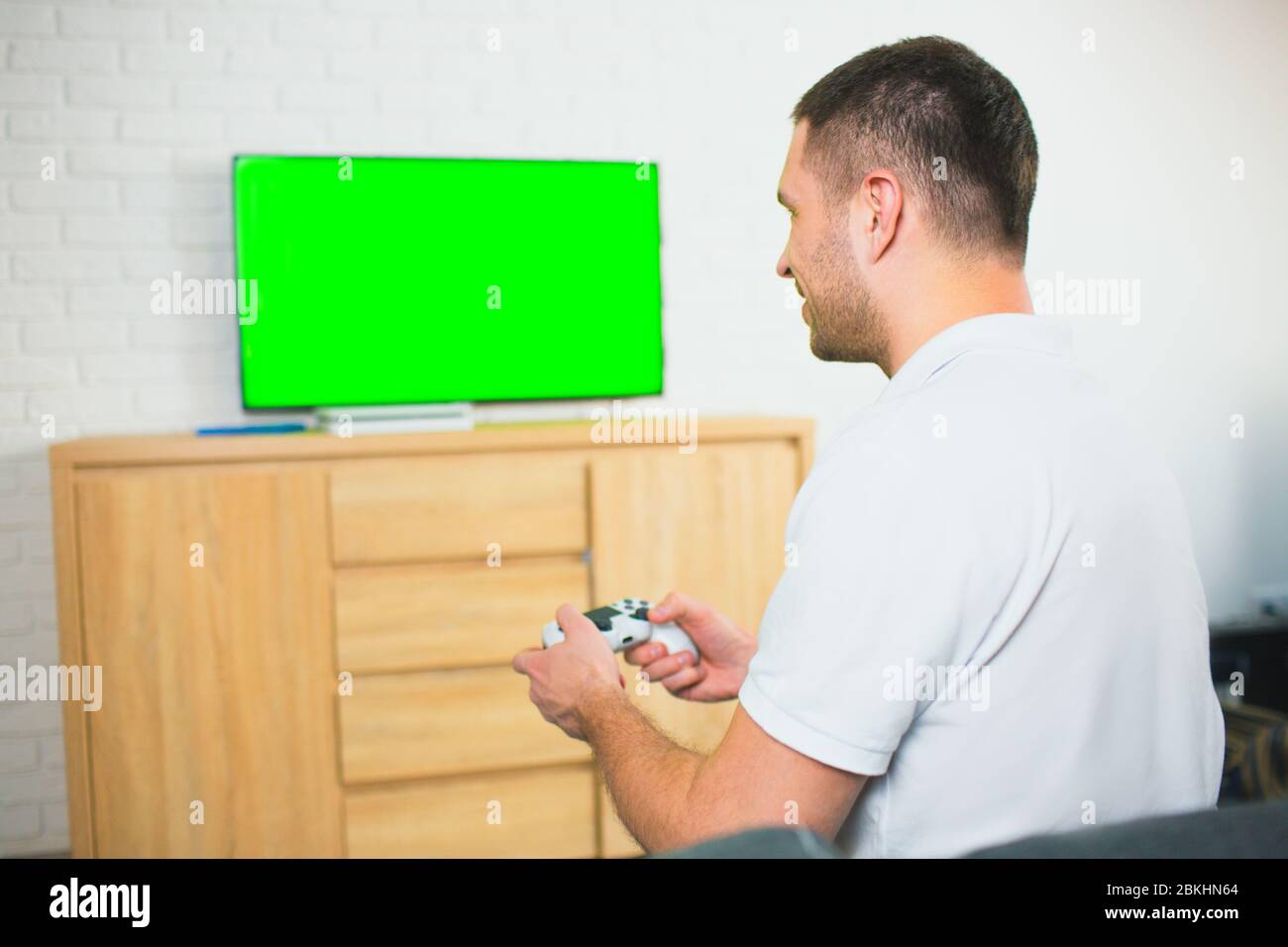 Junger Mann sitzt während der Quarantäne im Zimmer. Guy Spiele allein mit Joystick oder Gamepad. Grüner Bildschirm auf dem fernseher. Spielvergnügen. Stockfoto