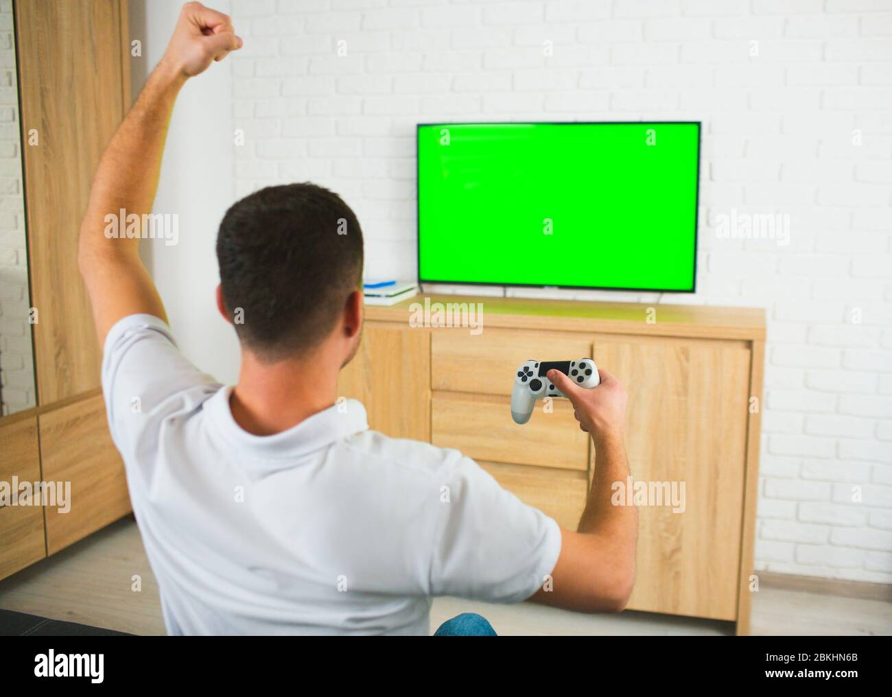 Junger Mann sitzt während der Quarantäne im Zimmer. Guy Computerspiele mit Joystick und tv. Grüner Bildschirm. Alleine spielen und jubeln. Rückansicht. Stockfoto