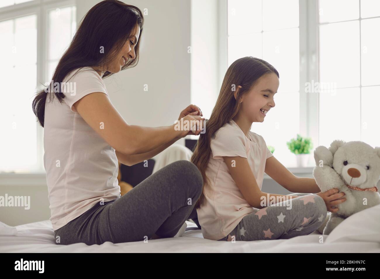 Mutter macht Frisur zu Mädchen Kind auf dem Bett im Schlafzimmer sitzen. Stockfoto