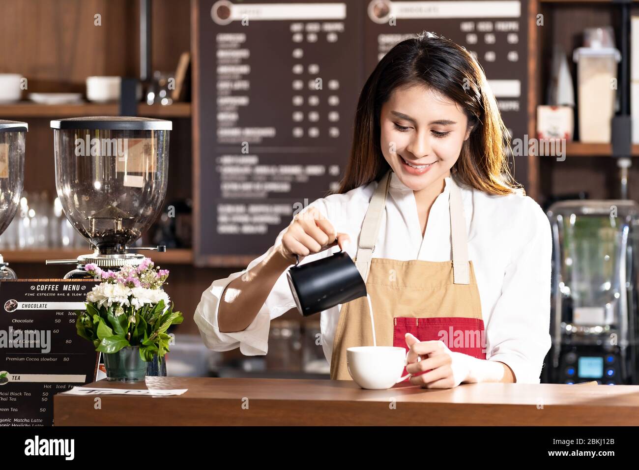 Junge Erwachsene asiatische Frau Barista Gießen frische Milch Latte Kaffee für den Kunden in Café-Bar mit ihrem Kollegen im Hintergrund zu bereiten. F Stockfoto