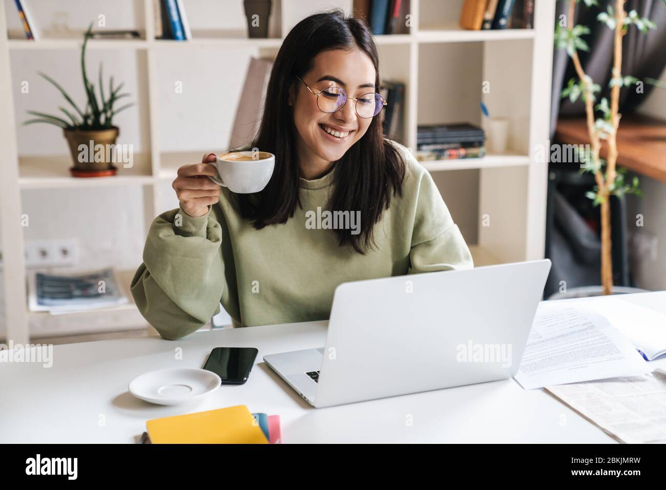 Attraktive glückliche junge Studentin, die an der Universitätsbibliothek studiert, am Schreibtisch sitzt, Laptop-Computer benutzt, Kaffee trinkt Stockfoto