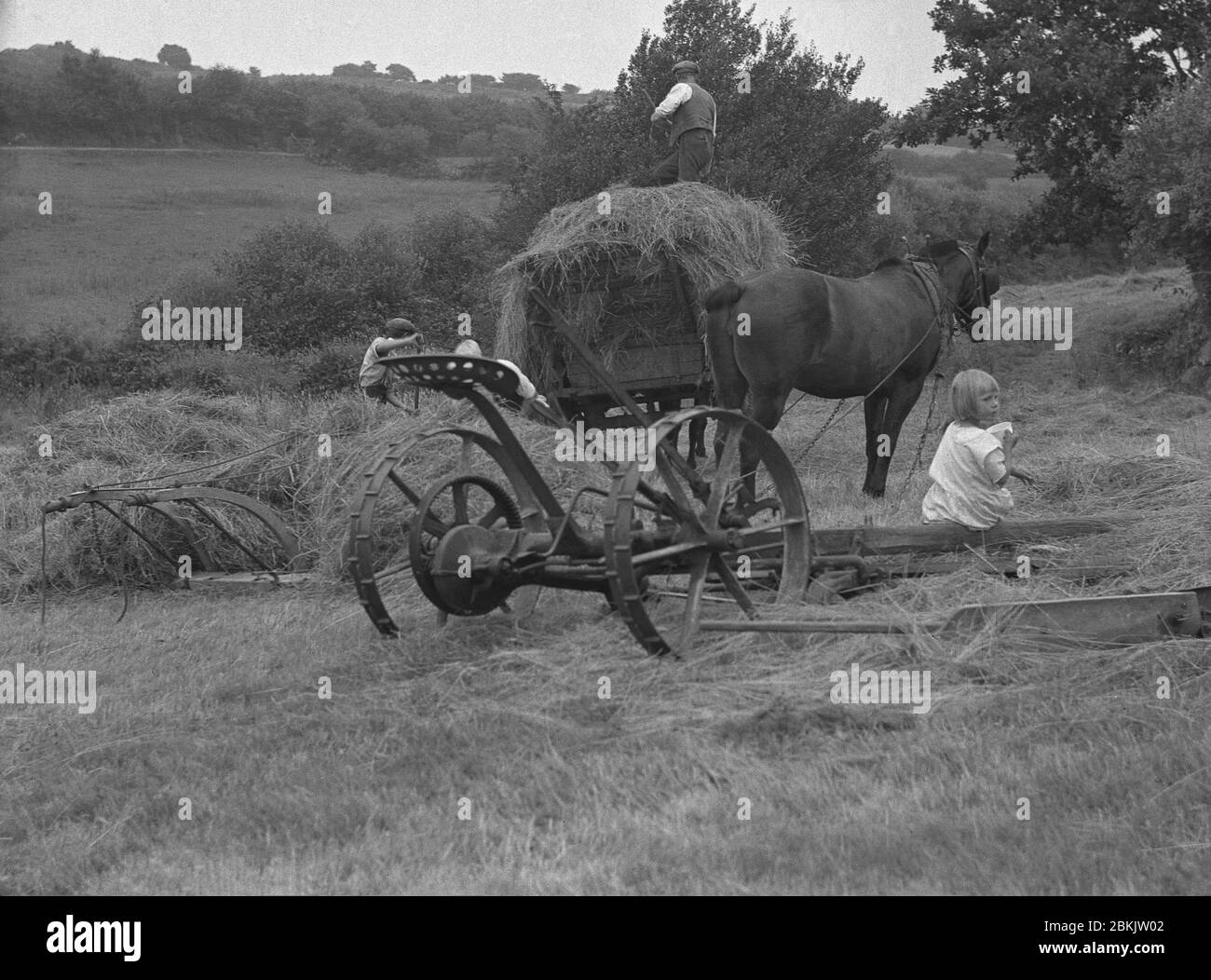 1930er Jahre, historisches Bild, in den Hügelketten bei Dartmoor, in Chagford, Devon, arbeiten zwei Bauern draußen auf einem Feld, sammeln Heu in einem Holzwagen, mit einem kleinen Mädchen, einer Bauernmädchen, die auf den Holzbeinen des zweisäckigen Anbaugerätes sitzt. In dieser Zeit war das Stapeln von Heu auf den Wagen zeitaufwendige, schwere körperliche Arbeit, aber wichtig für das Winterfutter für die Nutztiere des Hofes. Stockfoto