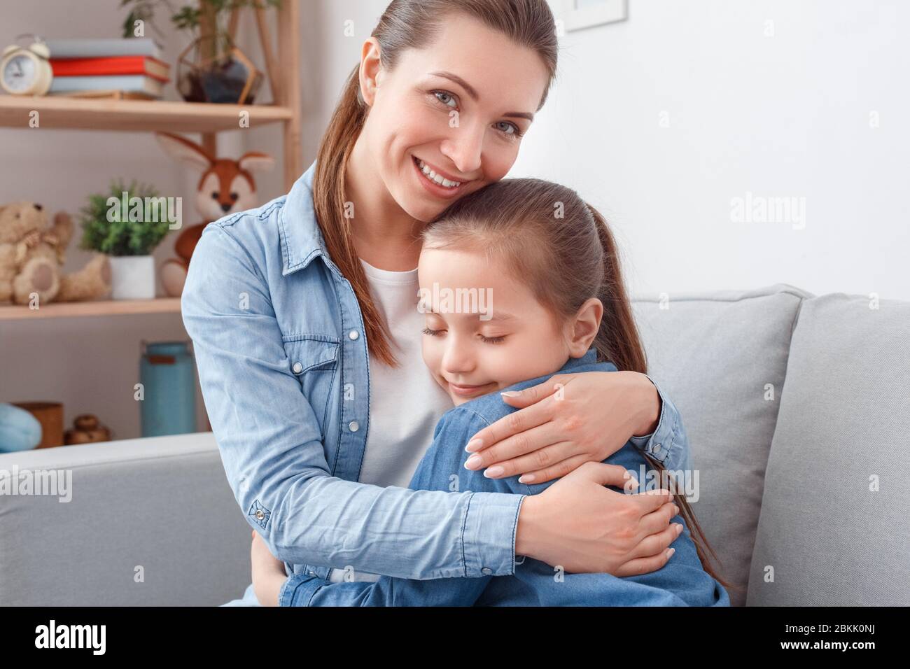 Lächelnde junge Erwachsene Frau umarmt ihre kleine Tochter sitzt auf der Couch Stockfoto