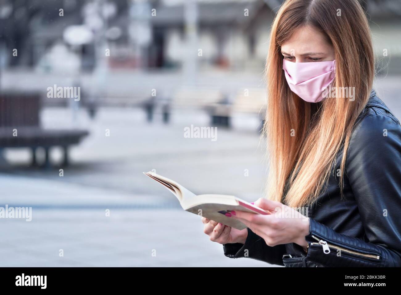 Junge Frau mit Hand gemacht rosa Gesicht Nase Mund Maske sitzen allein, Buch lesen, leere Stadtplatz hinter ihr. Kann während Coronavirus-Covid19 verwendet werden Stockfoto