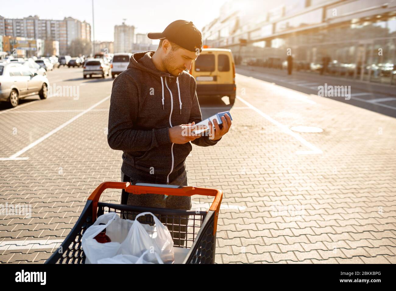 Ein Mann liest die Zusammensetzung der Lebensmittelzutaten auf der Verpackung auf dem Parkplatz eines Einkaufszentrums oder Supermarktes. In der Nähe steht ein Kinderwagen für Stockfoto