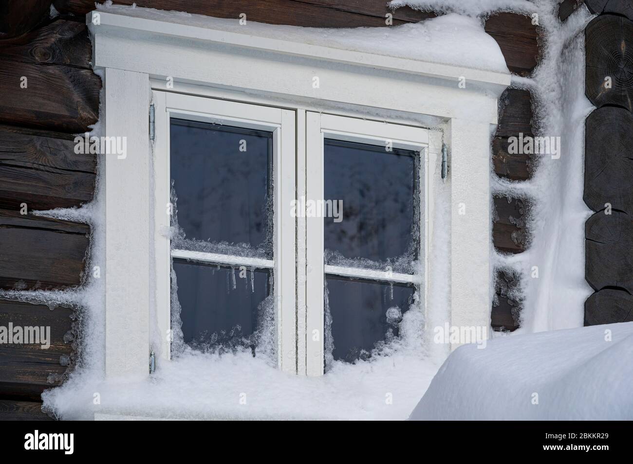 Weißer Fensterrahmen auf einem dunkelbraunen Holzhaus, das mit Schnee bedeckt ist Stockfoto
