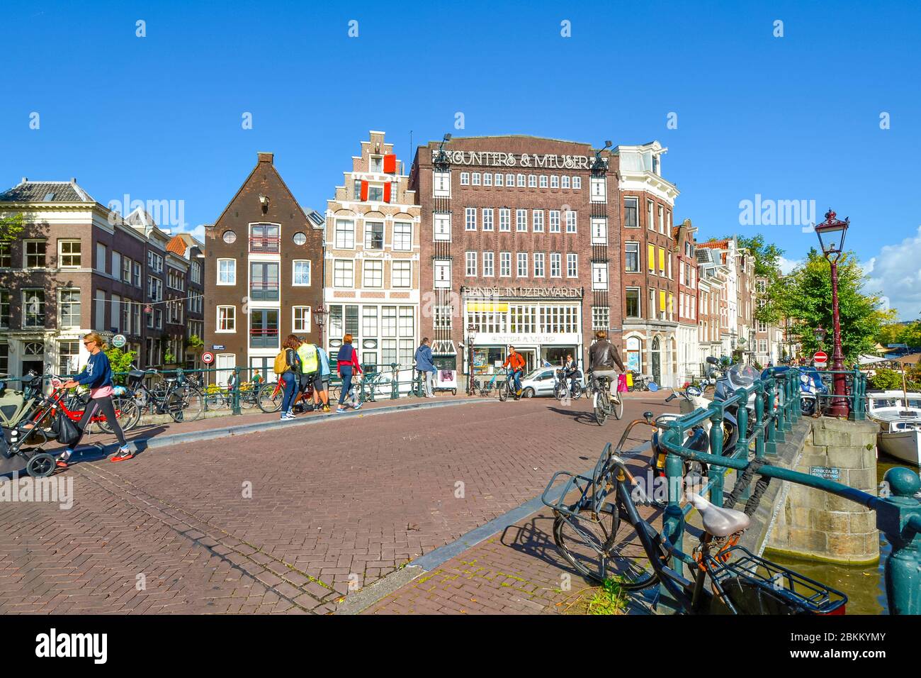 Fahrräder parken entlang eines Kanals in der Nähe der Westerkerk Kirche, während Touristen und Einheimische die Geschäfte und Brücken im historischen Zentrum von Amsterdam Nether genießen Stockfoto