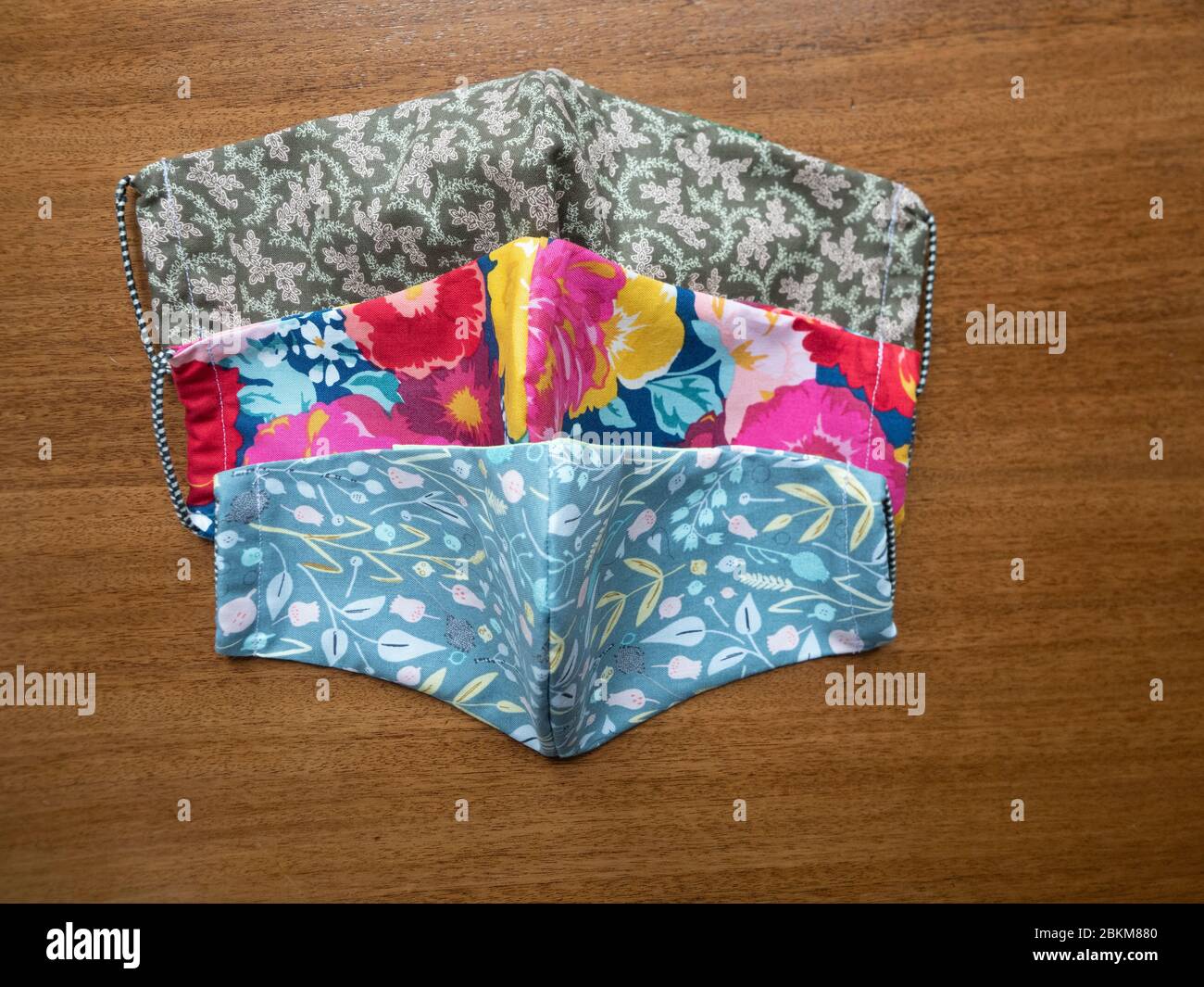 Drei handgefertigte Stoffmasken mit Blumenmuster auf einem Holztisch, fotografiert mit selektivem Fokus von oben. Stockfoto