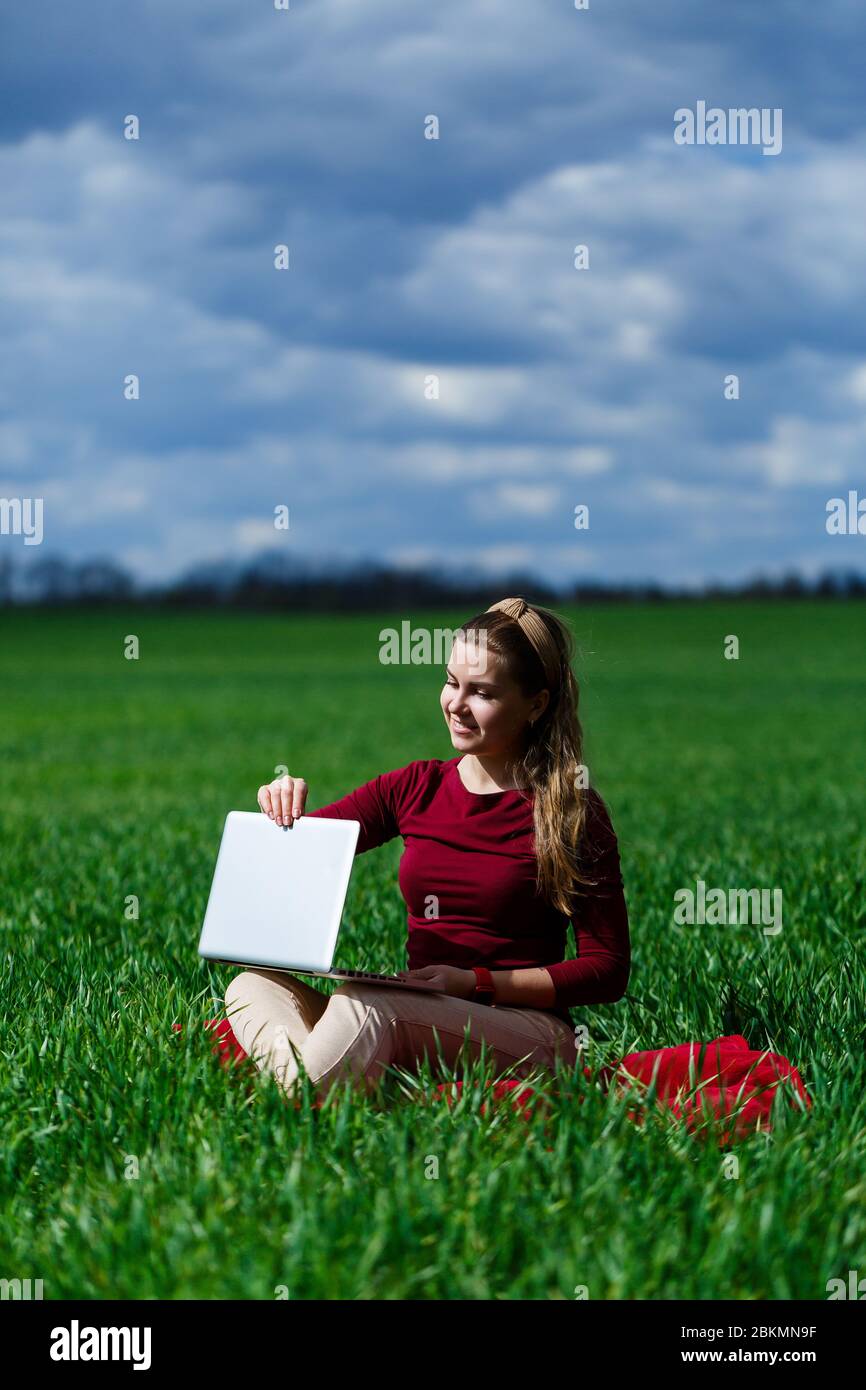 Fröhliches junges Mädchen auf dem Gras mit einem Laptop auf ihrem Schoß. Er hob die Hände hoch und lachte. Glück im Lebensstil eines klassischen Studenten. Arbeit o Stockfoto