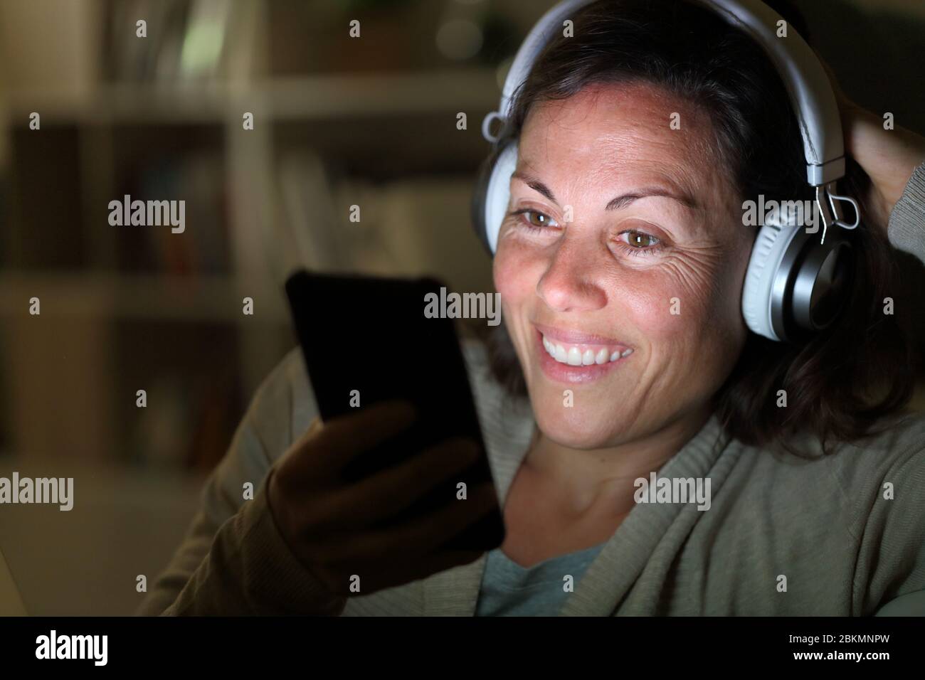 Glückliche Erwachsene Frau hört Musik mit Kopfhörern auf Smartphone mit beleuchteten Bildschirm sitzen in der Nacht zu Hause Stockfoto