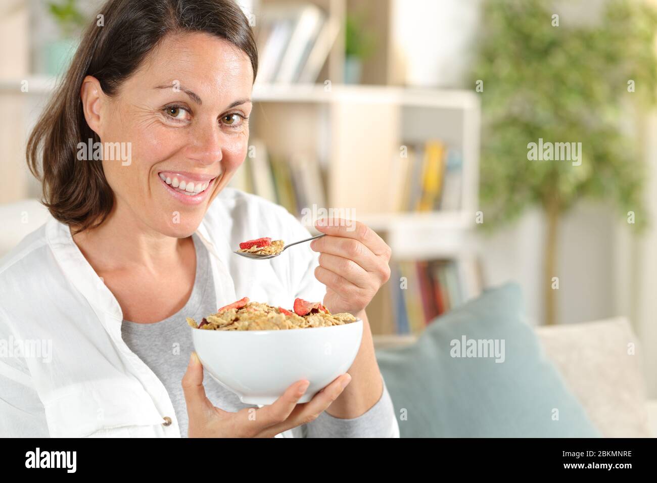 Glückliche Erwachsene Frau mit Müslischale mit Obst beim Frühstück sitzen auf dem Sofa zu Hause Stockfoto
