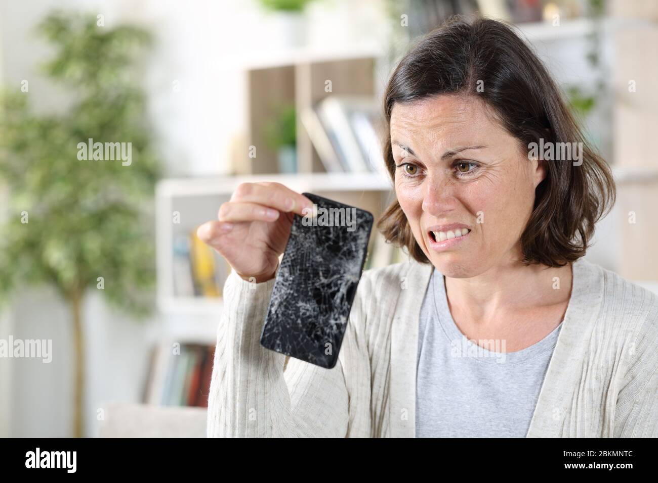 Verwirrt Erwachsene Frau Blick auf gebrochene Smartphone mit zerbrochenen Bildschirm zu Hause Stockfoto