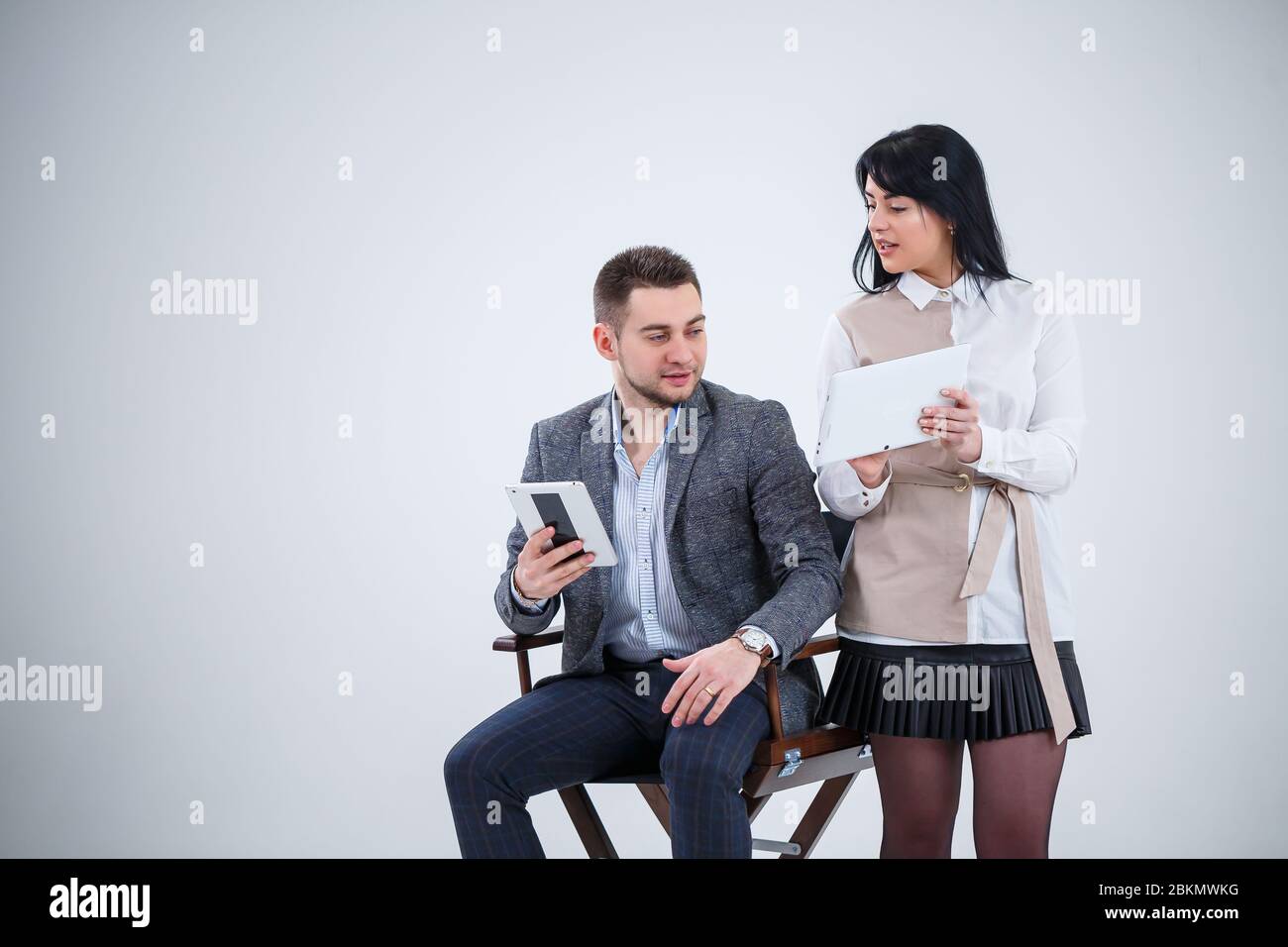 Ein erfolgreicher Mann in einem Anzug sitzt auf einem schwarzen Stuhl und eine Frau steht neben dem Blick auf die Tablets. Zukünftige Geschäftsleute lächeln und sprechen von einer ne Stockfoto