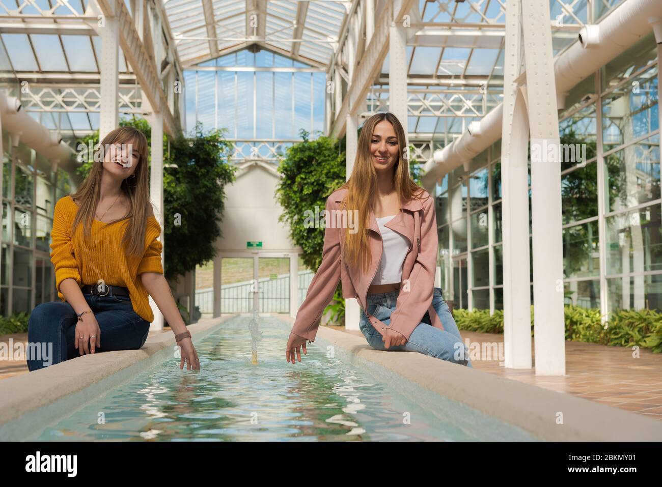 Zwei junge kaukasische Frauen sitzen gerne am Rande eines Brunnens mit Wasser in einem hellen Gewächshaus Stockfoto