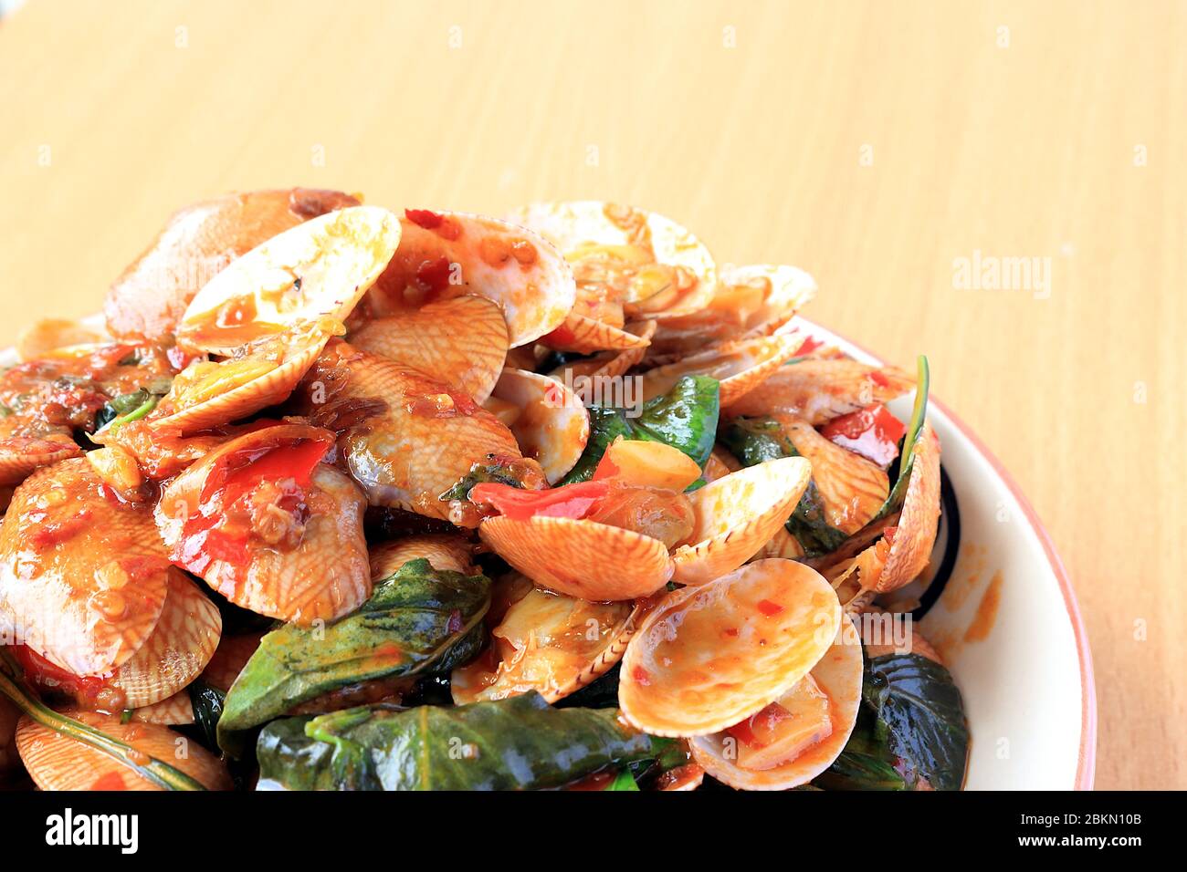 Gebratene gestreifte Muscheln mit Basilikum, Chilischote, Knoblauch und frischen Chilischoten verrühren. Platziert in einer weißen Platte mit einem schönen Muster und auf dem Holzmuster Stockfoto