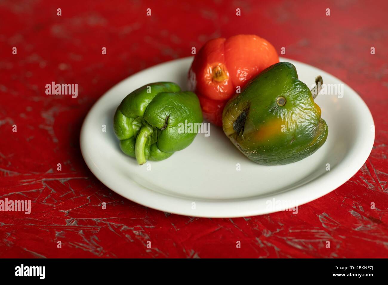 Verfaulte rote und grüne Paprika auf einem weißen Teller und einem roten Holzhintergrund. Lebensmittelabfälle, Kompost. Altes schimmeliges Gemüse, horizontal Stockfoto