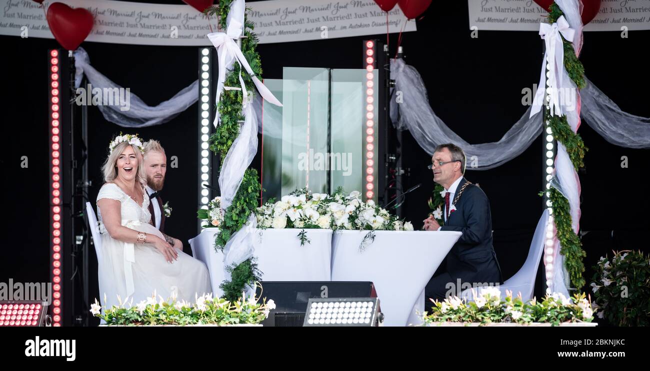 Düsseldorf, Deutschland. Mai 2020. Das Brautpaar Janine und Philip Scholz (l) sitzen mit Düsseldorfs Oberbürgermeister Thomas Geisel (SPD) bei ihrer Hochzeitszeremonie im Drive-in-Kino auf der Bühne. Drei Paare sagen sich im Drive-in-Kino Ja. Die Gäste können die Zeremonie im Autoradio hören. Das Paar heiratete ganz in Rot und Weiß - zu Ehren des 125-jährigen Bestehens des Fußballvereins Fortuna Düsseldorf. Quelle: Fabian Strauch/dpa/Alamy Live News Stockfoto