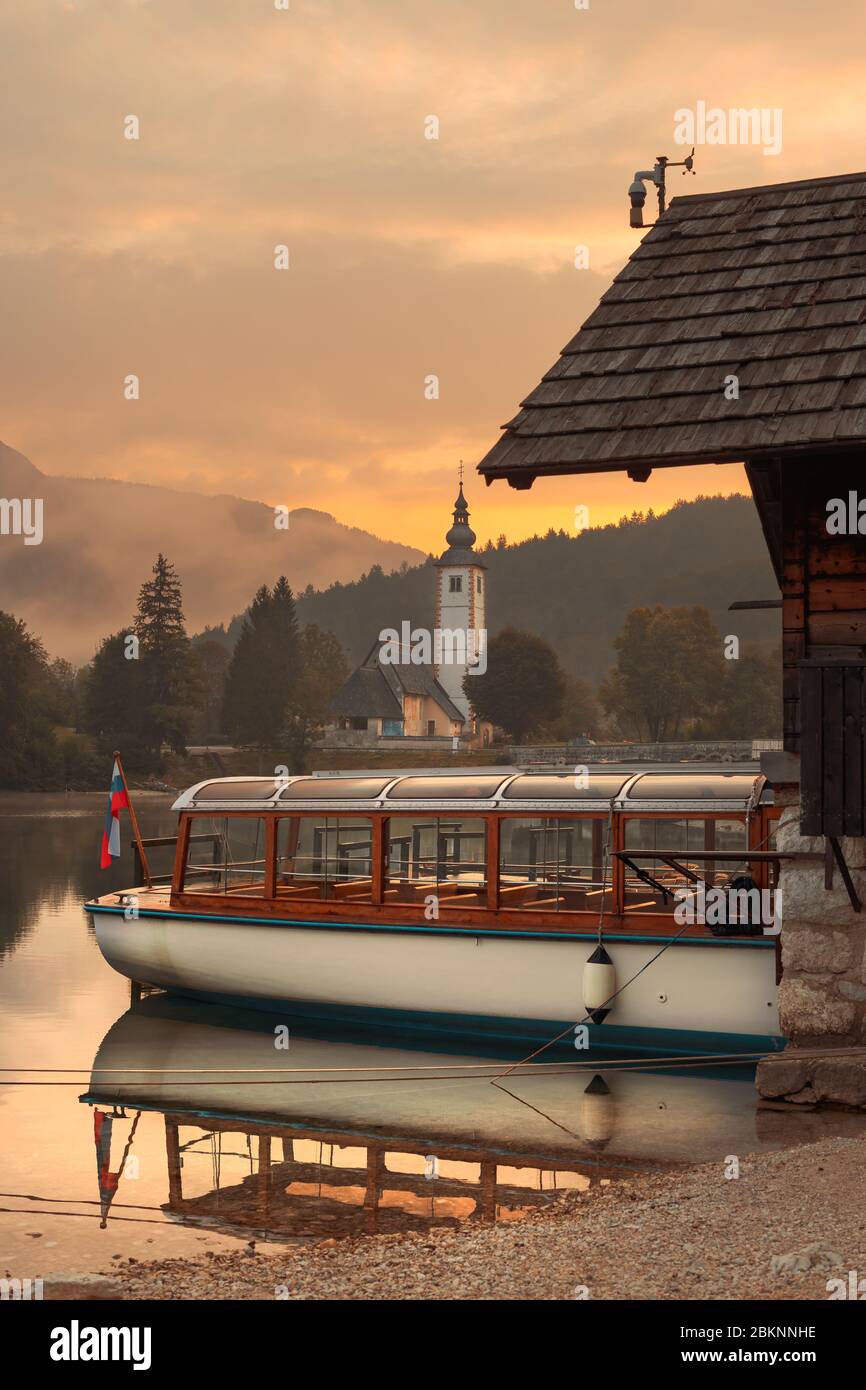 Elektrisches Motorboot für die Besichtigung des Bohinjer Sees in Slowenien, Kirche des heiligen Johannes des Täufers und alte Steinbrücke im Hintergrund Stockfoto