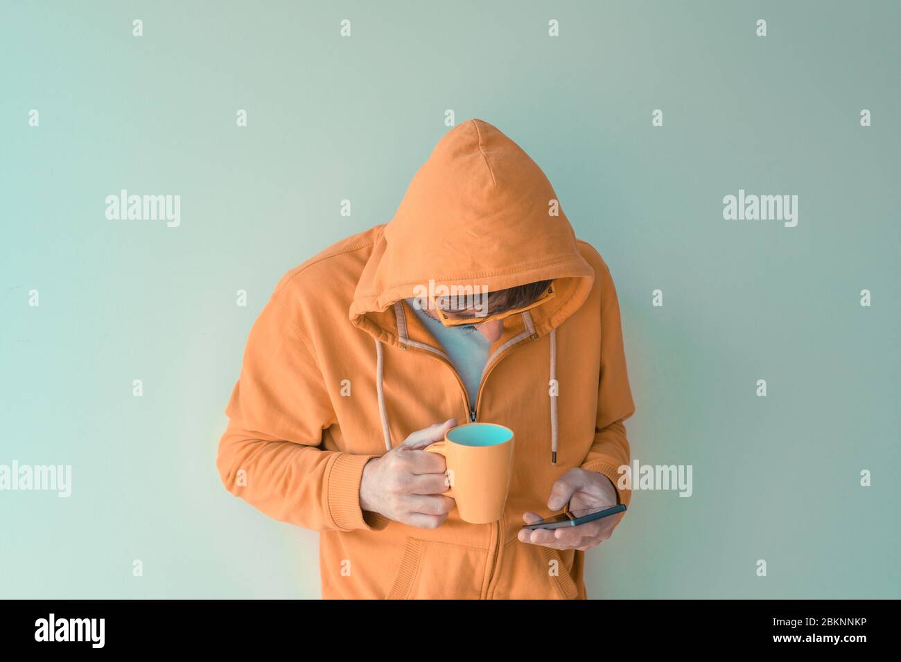 Mann trinkt Kaffee und benutzt morgens das Handy, Erwachsener Mann mit gelber Kapuze mit Reißverschluss für Kaffeetasse und Smartphone, selektive Fokussierung Stockfoto