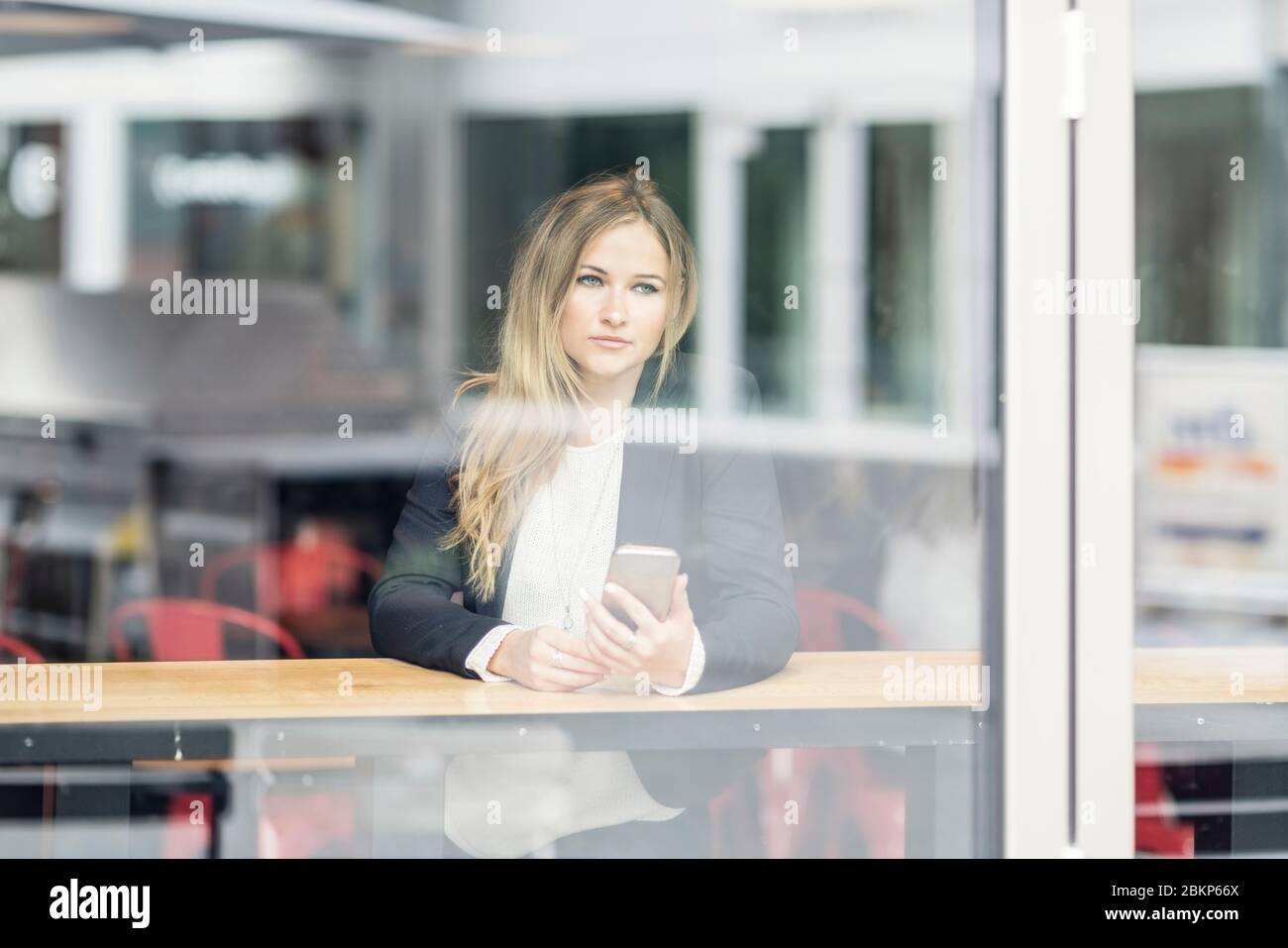 Lächelnde blonde Frau mit Smartphone, die in einem Café in Berlin an einem Fenster sitzt. Stockfoto