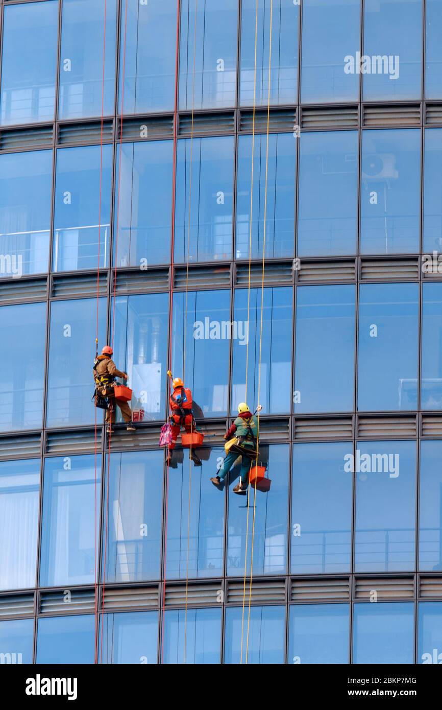 Drei Männer Arbeiter in roten und dunklen Arbeitskleidung reinigen die Außenfenster eines Business-Wolkenkratzers. Im Freien Stockfoto