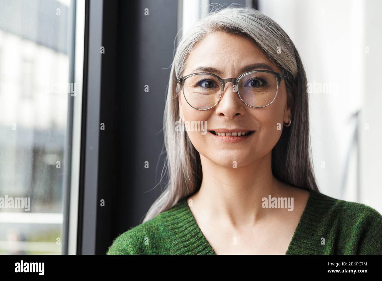 Attraktive lächelnde reife Frau mit langen grauen Haaren, die drinnen am Fenster stehen und die Kamera betrachten Stockfoto