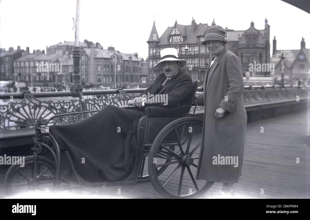 Ende der 1920er Jahre, historisch, auf einem Gehweg an einer viktorianischen Küste, ein Herr in einem Hut sitzt in einem Einsitzer dreirädrigen ungültige Wagen der Zeit, mit einer Decke über seinen Beinen und seine Frau neben ihm, England, UK. Diese Mobilitätsfahrzeuge hatten eine Deichsel, die mit dem Vorderrad für die Lenkung verbunden war. Stockfoto