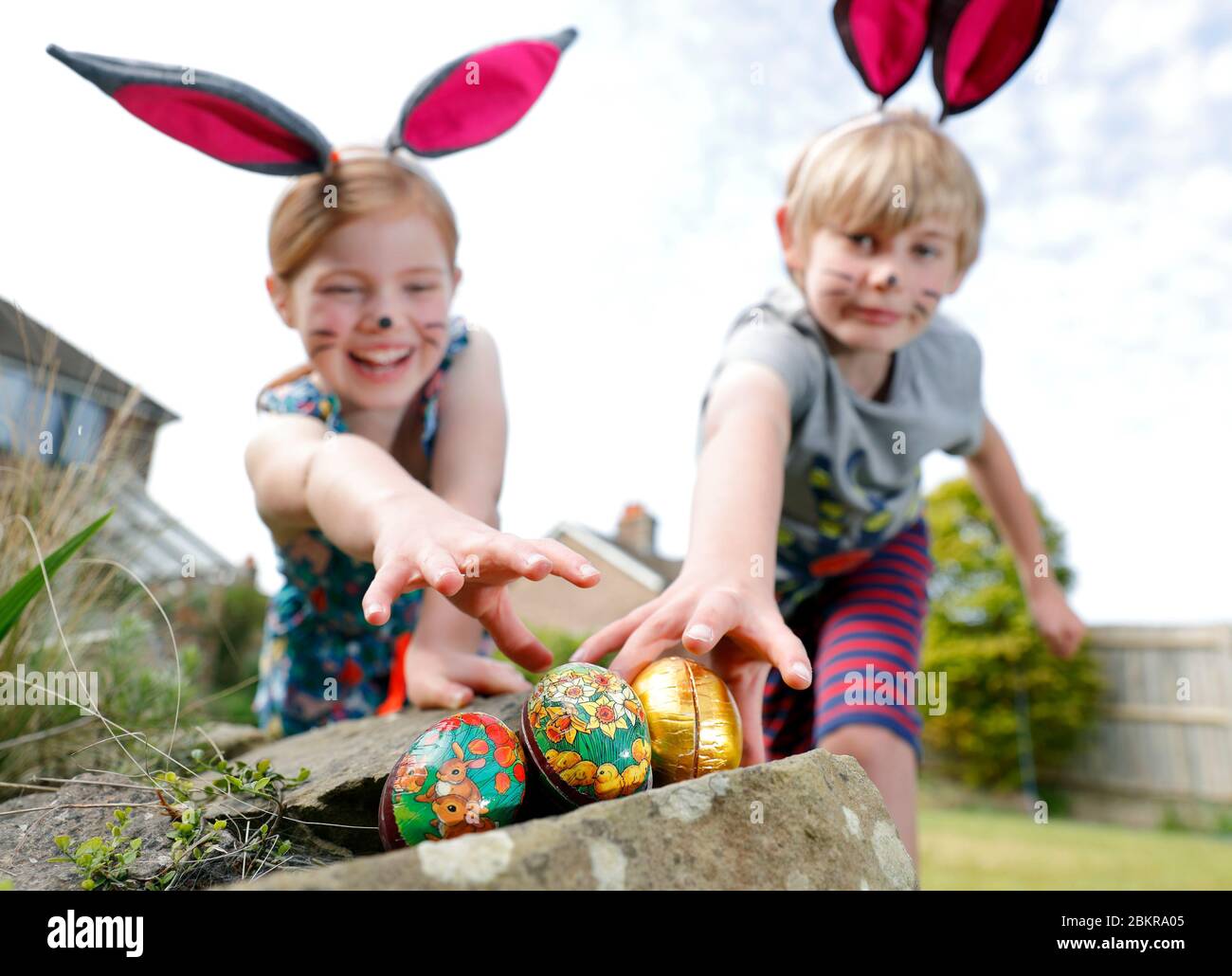 Ein Bruder und eine Schwester, die Gesichtsfarbe und hausgemachte Hasenohren tragen, nehmen an einer Ostereiersuche am Ostersonntag in ihrem Garten Teil. Stockfoto