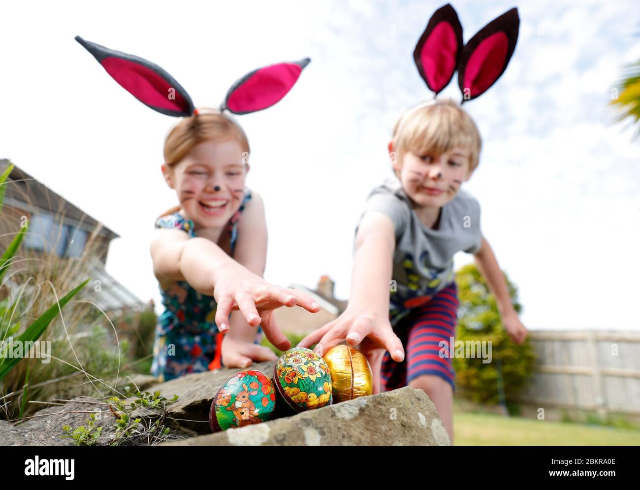 Ein Bruder und eine Schwester, die Gesichtsfarbe und hausgemachte Hasenohren tragen, nehmen an einer Ostereiersuche am Ostersonntag in ihrem Garten Teil. Stockfoto