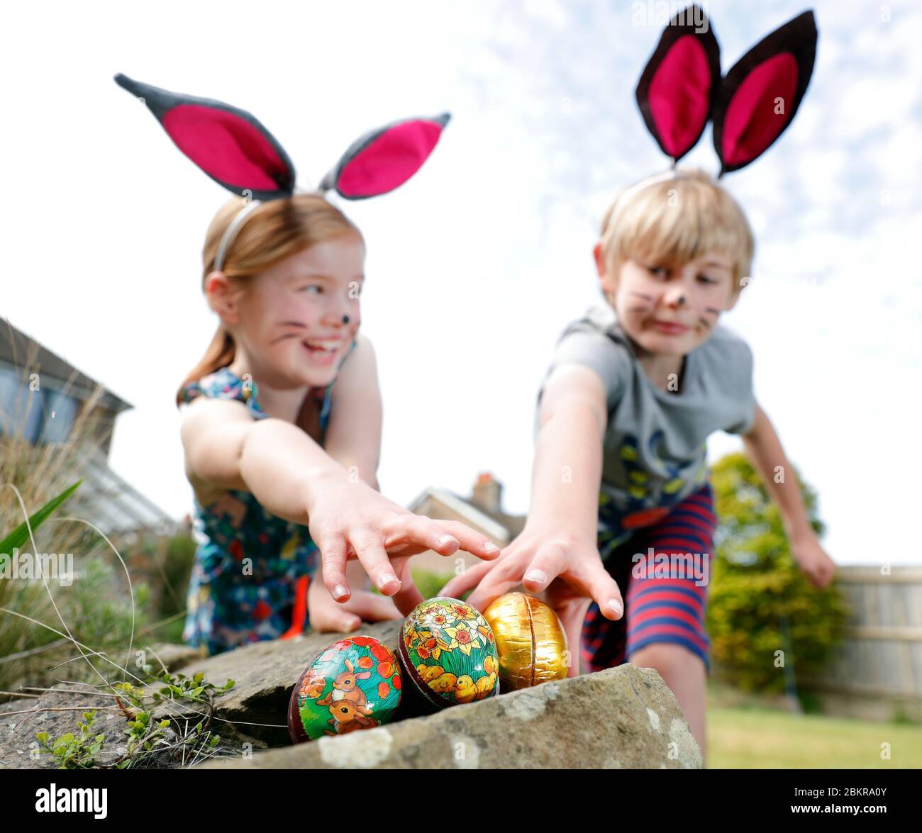Ein Bruder und eine Schwester, die Gesichtsfarbe und hausgemachte Hasenohren tragen, nehmen an einer Ostereiersuche am Ostersonntag in ihrem Garten Teil. Stockfoto