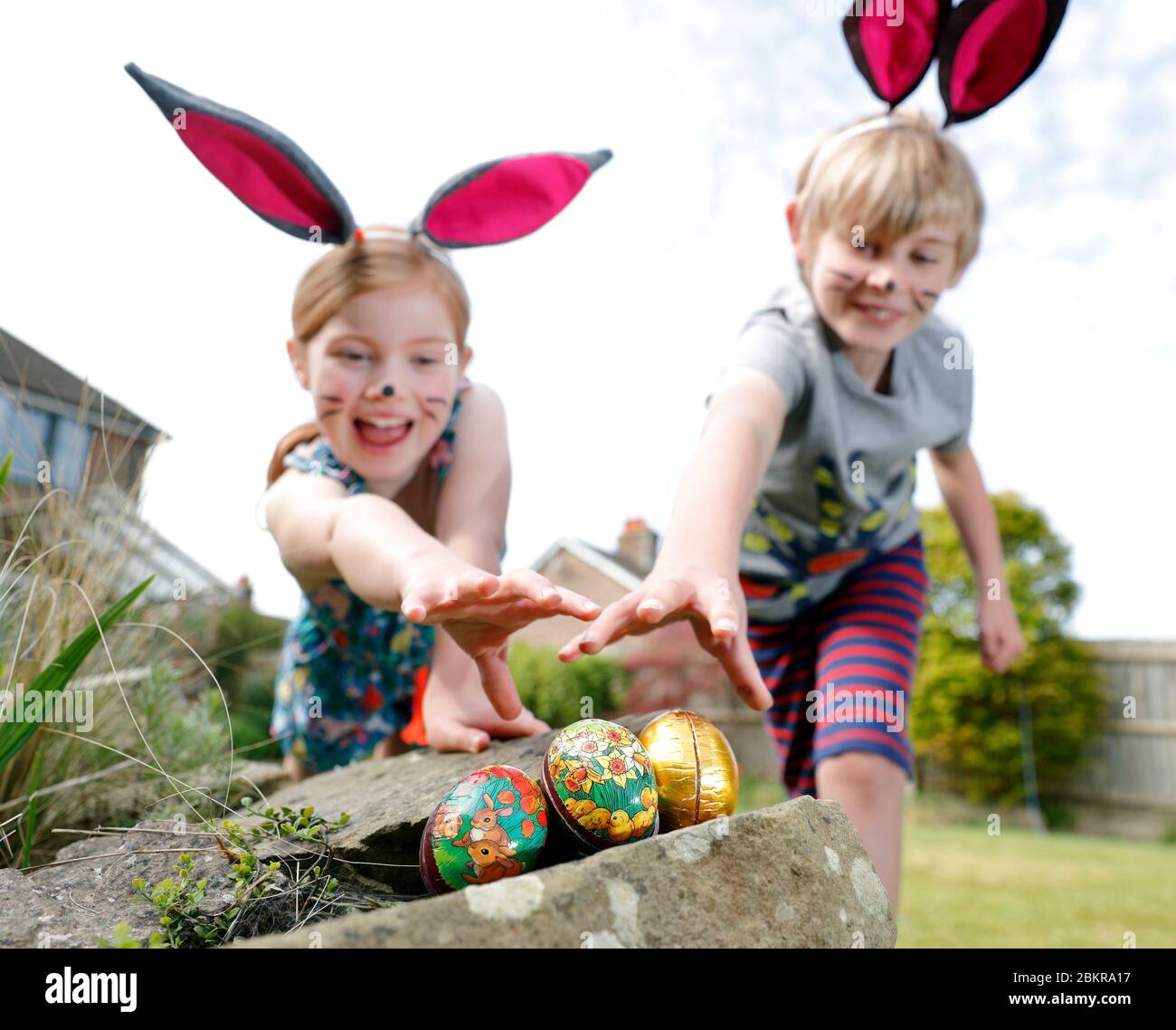 Ein Bruder und eine Schwester, die Gesichtsfarbe und hausgemachte Hasenohren tragen, nehmen an einer Ostereiersuche am Ostersonntag in ihrem Garten Teil. Stockfoto
