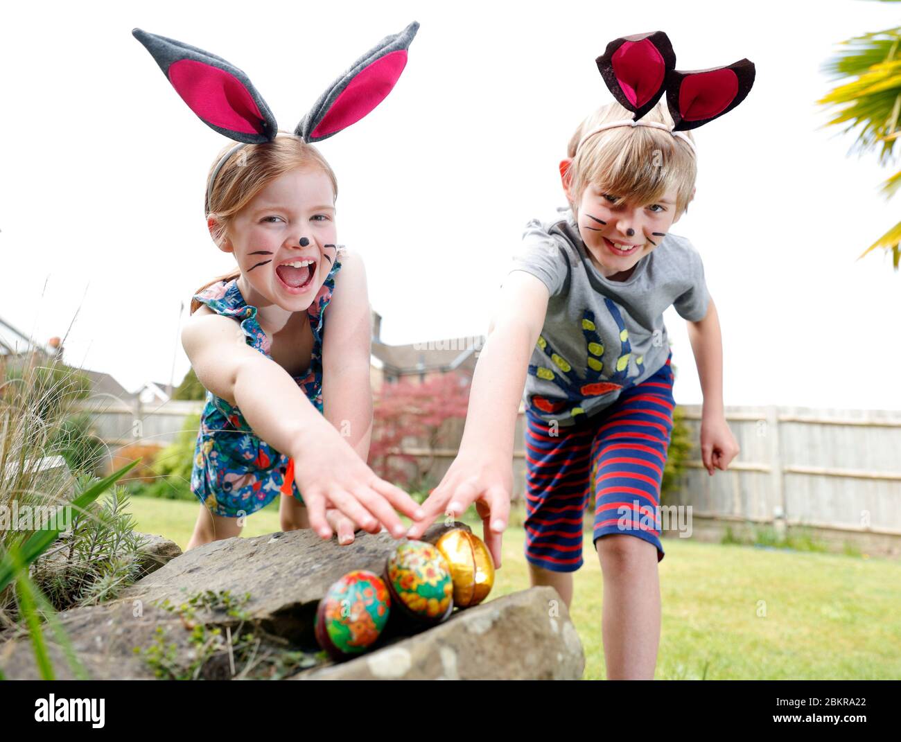 Ein Bruder und eine Schwester, die Gesichtsfarbe und hausgemachte Hasenohren tragen, nehmen an einer Ostereiersuche am Ostersonntag in ihrem Garten Teil. Stockfoto