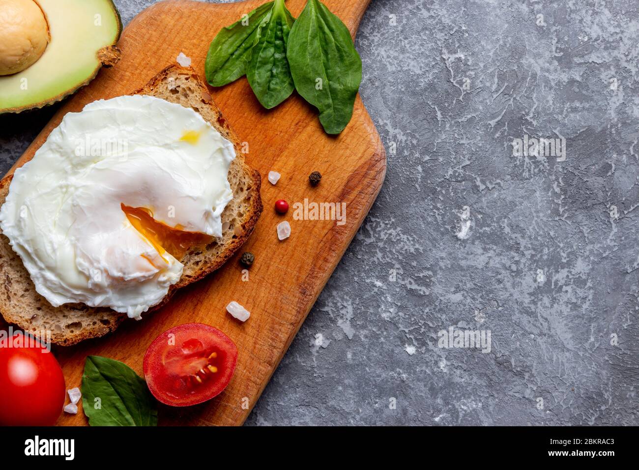 Pochiertes Ei auf Brot mit Samen, Kirschtomate, Spinat, Avocado, Salz und Gewürzen auf einem Holztablett auf Steingrund Stockfoto