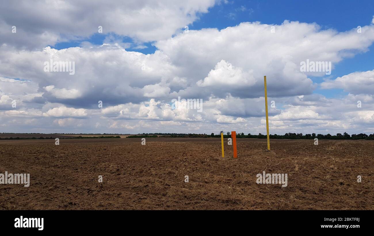 Dramatischer Himmel über der ländlichen Landschaft des braunen Landfeldes. Metallstangen in rot und gelb lackiert. Wunderschöne Wolkenlandschaft mit weißen Cumulus-Wolken. Stockfoto