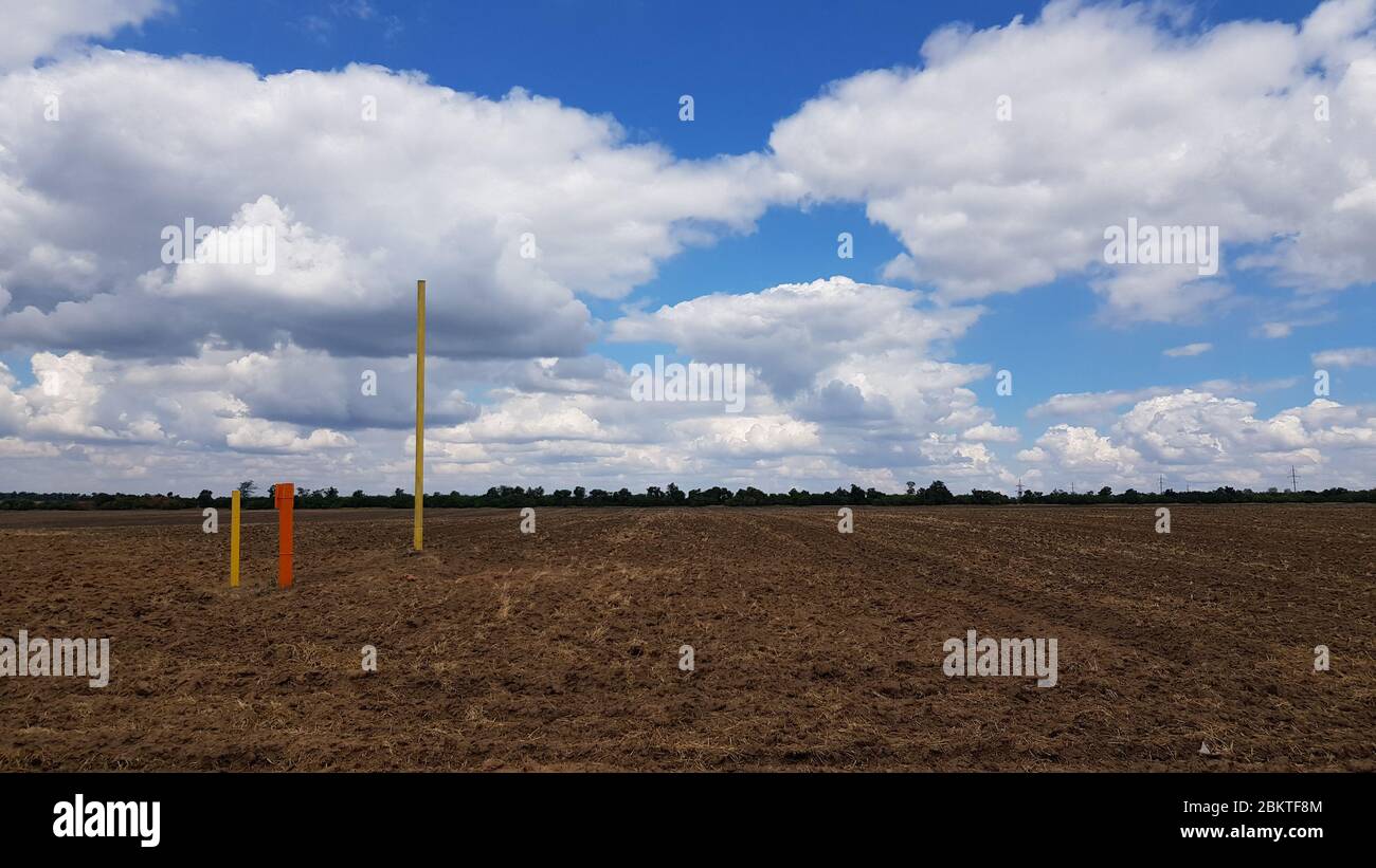 Landwirtschaftliche Landschaft von braunen Erdeggen und hohen blauen Himmel mit flauschigen Wolken. Metallstangen in rot und gelb lackiert. Wunderschöne Wolkenlandschaft Stockfoto