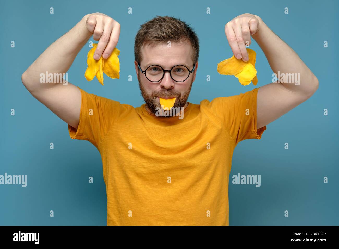Verspielter, schelmiger Mann in runden Gläsern und einem gelben T-Shirt hält trockene Mango in Mund und Händen auf blauem Hintergrund. Stockfoto