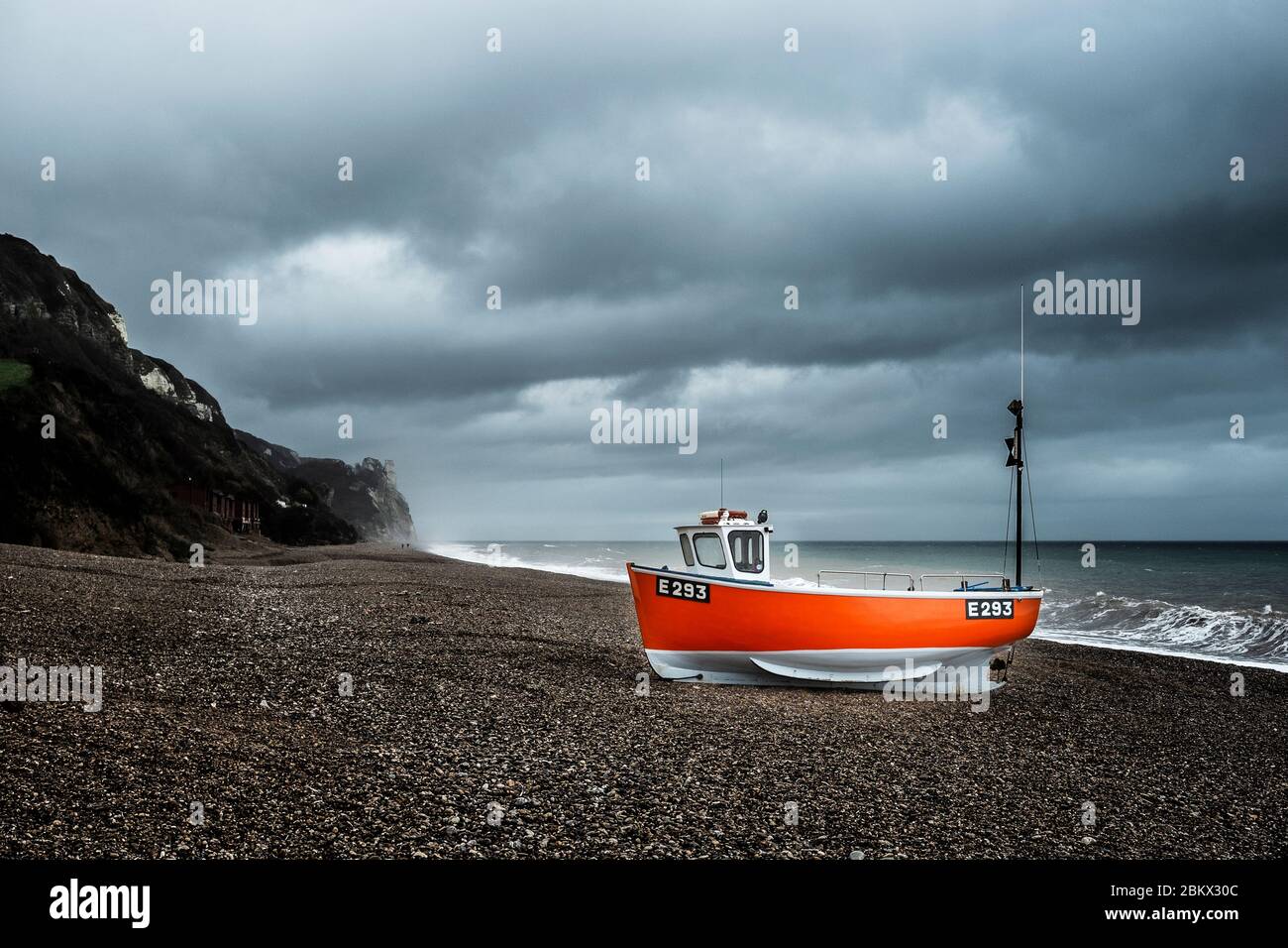 Ein rot gehultes Fischerboot am Kiesstrand von Branscombe, Devon mit stürmischem Himmel Stockfoto
