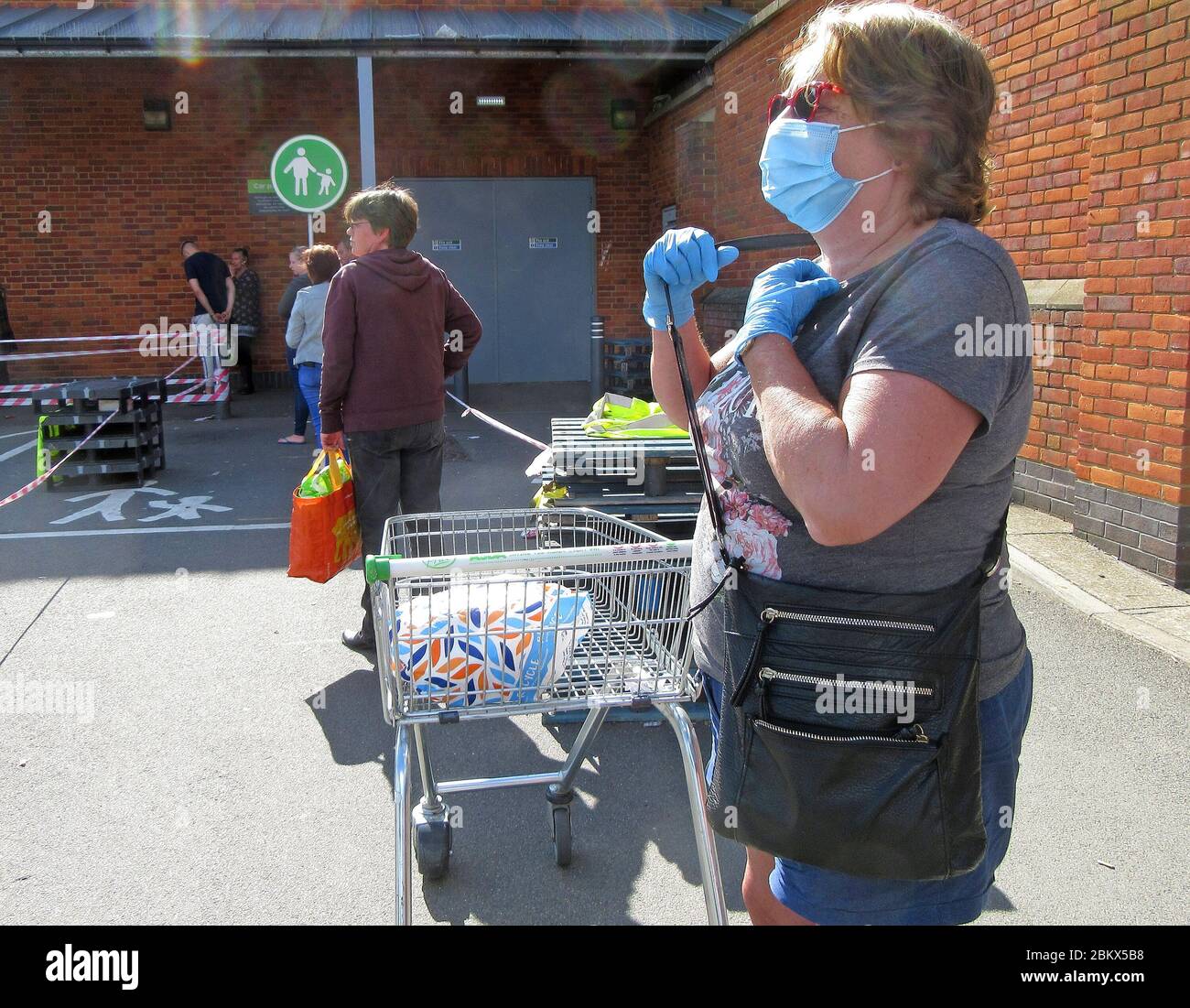Eine Frau, die eine Gesichtsmaske und Handschuhe als Vorsichtsmaßnahme trägt, während sie in einer Warteschlange in einen Supermarkt inmitten der Coronavirus-Pandemie eingeht.Shopper Schlange stehen und beachten Sie die notwendige soziale Distanz, die in Großbritannien zur neuen Regel geworden ist. Stockfoto