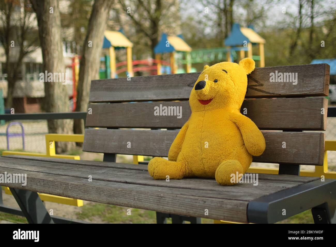 Gelbbär Spielzeug sitzt auf der Bank in der Nähe von leeren geschlossenen Spielplatz im Freien ohne Kinder wegen Quarantänemaßnahmen gegen COVID-19 Stockfoto