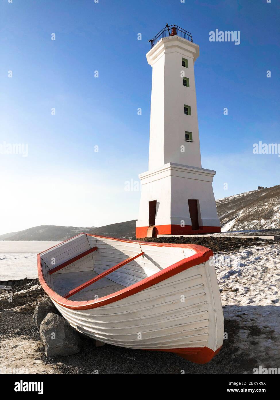 Bild von altem Leuchtturm und Holzboot an der Küste von Magadan, Russland. Hohes weißes Gebäude und Boot mit roten Streifen. Blauer Himmel und Eis auf dem Meer im Winter. Stockfoto