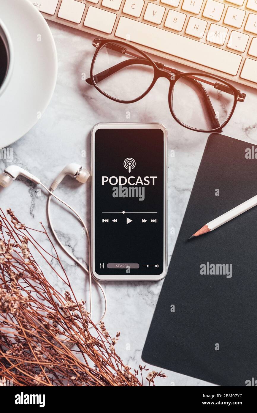 Podcast Audio Content Konzept. Podcast-Anwendung auf mobilen Smartphone-Bildschirm auf Arbeitsplatz Schreibtisch mit Kaffeetasse, Kopfhörer, Brille, Computer-Keyboar Stockfoto