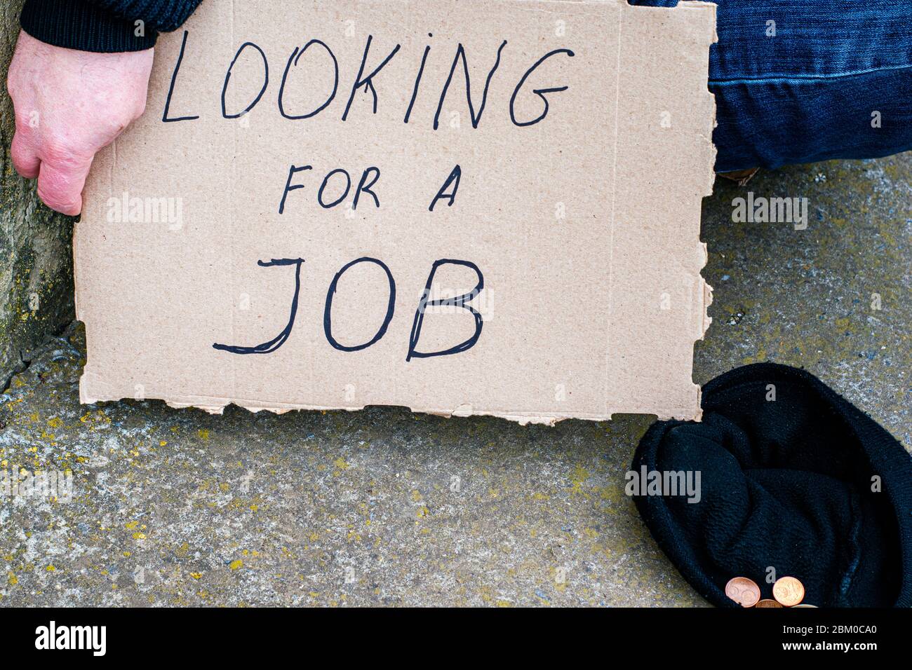 Arbeitsloser Mann sitzt auf dem Boden mit einem Karton Schild sagen, auf der Suche nach einem Job neben ist ein Hut mit Almosen Münzen Stockfoto