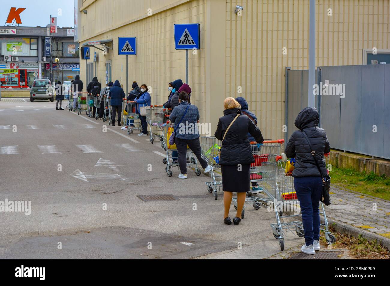 Schlange von Menschen mit Einkaufswagen auf dem Parkplatz des Lebensmittelladens. Menschen, die im Rahmen der Coronavirus-Beschränkungen wenige Meter voneinander entfernt stehen. Stockfoto