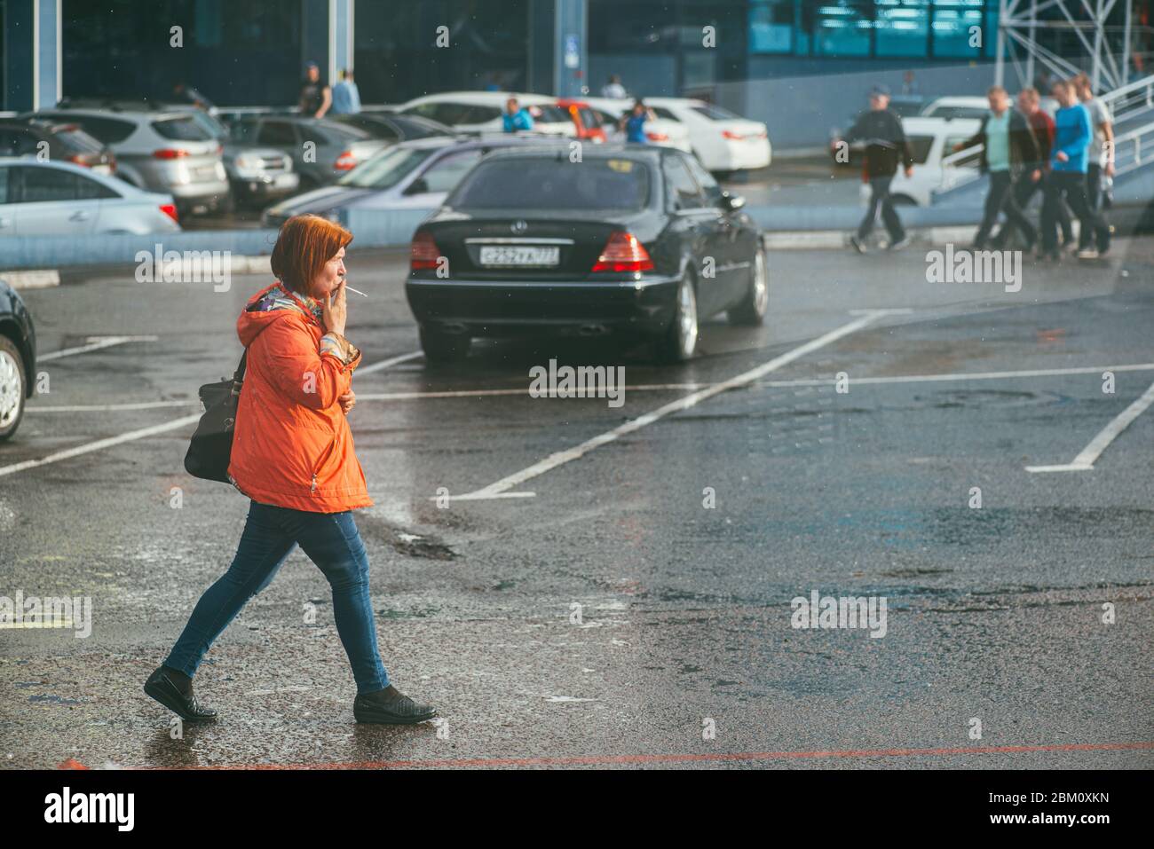 Moskau, Russland - 2. MAI 2018: Eine Frau in einer roten Jacke geht eine nasse Straße entlang und raucht an einem öffentlichen Ort eine Zigarette Stockfoto