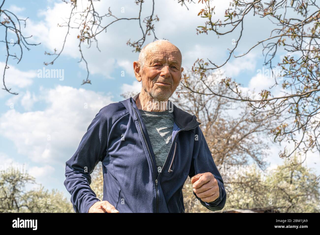 90 Jahre alter Mann in Sportbekleidung, der während des Frühlings sonniger Tag im blühenden Garten läuft Stockfoto