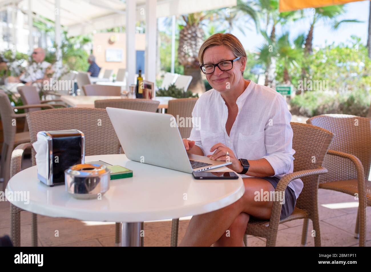 Frau sitzt in einer Kaffeebar und arbeitet an ihrem Laptop. Sie schaut mit einem glücklichen Lächeln in die Kamera. Stockfoto