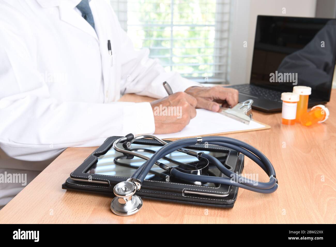 Nahaufnahme eines Stethoskops auf einem Medical Tablet Computer auf dem Schreibtisch mit Fenster und Arzt im Hintergrund auf einem Patientendiagramm. Der Schwerpunkt liegt auf dem Stockfoto