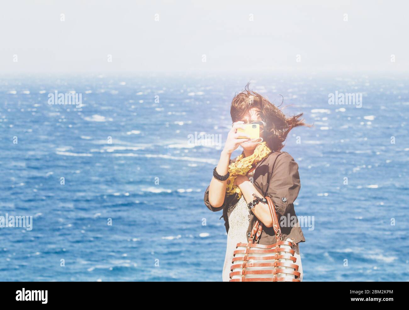 Porträt einer Modefrau gegen blauen Ozean an sonnigen Tag. Trendige junge Reisende in Freizeitkleidung machen ein Foto mit ihrer gelben Digitalkamera Stockfoto