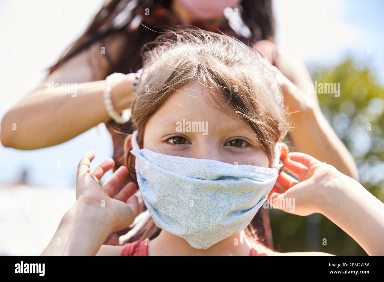 Eine Mutter bindet liebevoll eine Gesichtsmaske an ihre liebenswerte Tochter im Freien in einem Park, um eine Infektion durch Coronavirus zu verhindern Stockfoto